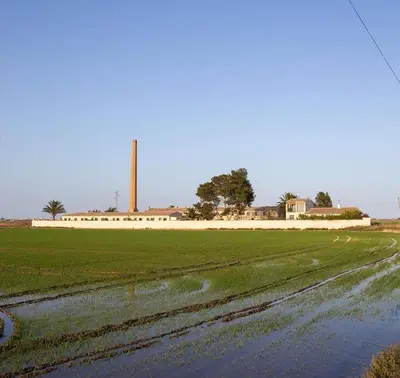 a field with a factory in the background and a dirt road at Hotel Rural El Teular in Sueca