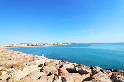 a man sitting on a rocky shoreline near the water at Global Vacacional, Bonito apartamento cerca de la playa in Puerto de Sagunto